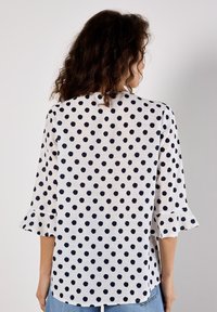 Weiße Bluse mit marineblauen Punkten, ausgestattet mit Dreiviertel-weit ausgestellten Ärmeln und einem lockeren Schnitt. Strukturiertem Stoff sorgt für visuelles Interesse.