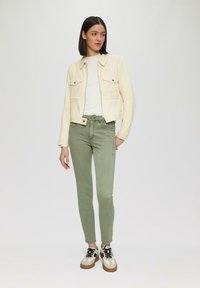 Vrouw met donker haar, gekleed in een crèmekleurige jas, witte top, groene skinny broek en beige sneakers, staande tegen een effen lichte achtergrond.