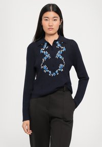 Námořnická polo svetr s dlouhým rukávem featuring květinovou výšivkou v modré a bílé barvě, žebrovanou texturou a dvouknoflíkovou légou. V kombinaci s tmavými kalhotami.