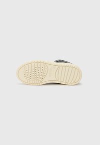 Semelle de sneaker avec un fond en caoutchouc blanc, texturé avec des rainures et des perforations pour une meilleure adhérence et ventilation ; tige noire avec des accents argentés.
