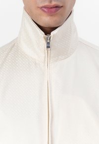 Ljusfärgad zip-jacka med hög krage, som har ett subtilt geometriskt mönster och slät textur, och visar upp silverdetaljer på dragkedjan.