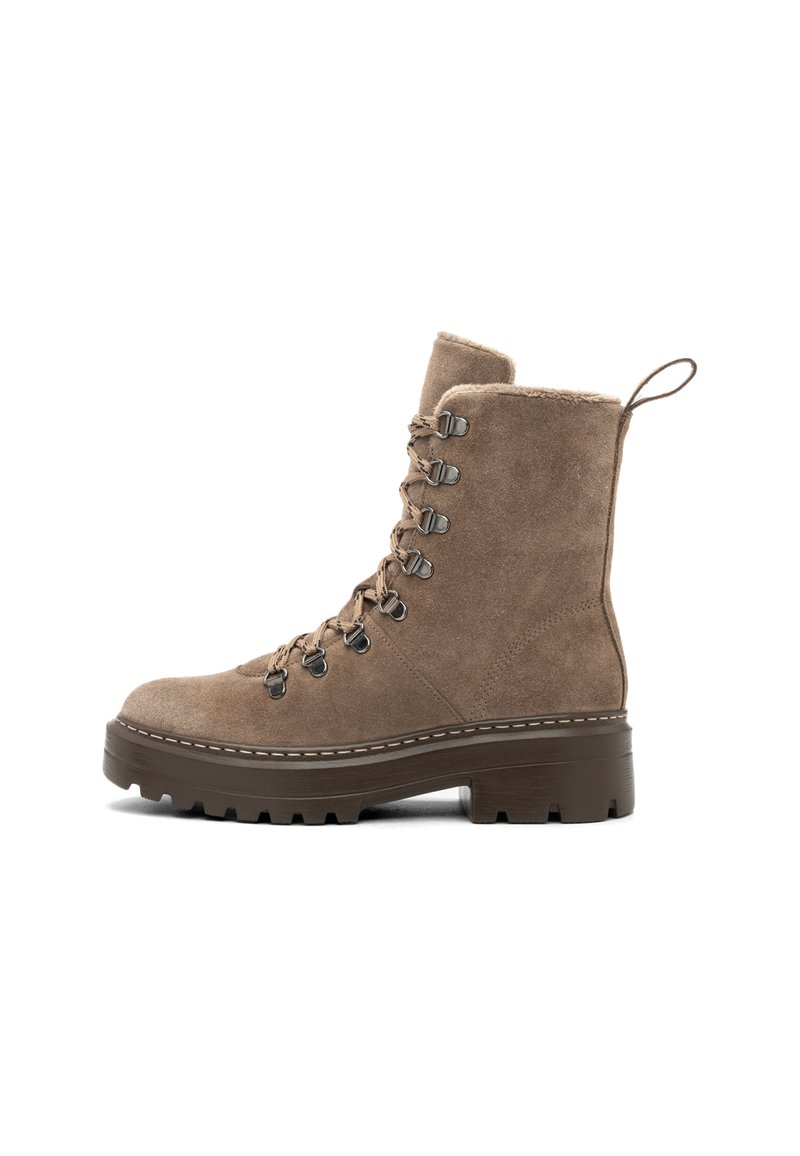 Bota de tobillo de ante marrón con cordones, suela gruesa y texturada, y forro de piel de oveja visible en el borde superior y la lengüeta.