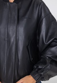 Schwarze Lederbomberjacke mit einem Reißverschluss vorne, geripptem Kragen und elastischen Bündchen, sowie einem gestickten Akzent in der Brustgegend.