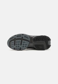 Semelle d'une chaussure de sport noire montrant des motifs détaillés de la bande de roulement avec des parties en caoutchouc noir et gris sur fond blanc.