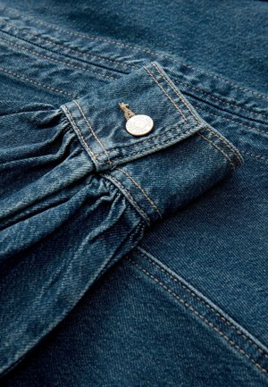 Nærbillede af en manchet på en blå denimjakke med samlet stof og en metaltrykknaplukning mod en matchende denim baggrund.