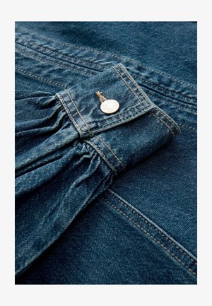 Nærbillede af en manchet på en blå denimjakke med samlet stof og en metaltrykknaplukning mod en matchende denim baggrund.