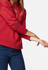 Rotes Sweatshirt mit Rundhalsausschnitt, verziert mit kleinen reflektierenden Akzenten, mit langen Ärmeln und lässigem Schnitt, kombiniert mit dunklen Jeans.