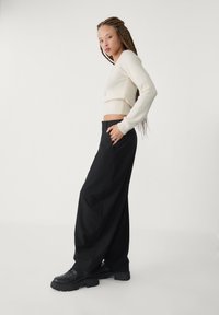 Pull court en crème avec des poignets côtelés, associé à un pantalon large noir taille haute et des bottes noires à lacets, présentant un tissu texturé et des poches.