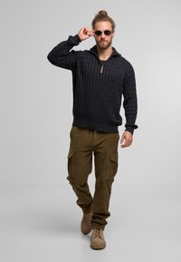Sort kabelstrikket sweater med en halv lynlås krave, parret med olivengrønne cargo bukser og beige snørestøvler. Model med solbriller.