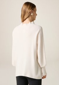 Kremowy, prążkowany sweter z wysokim kołnierzem i opuszczonymi szwami na ramionach, zestawiony z czarnymi spodniami o luźnym kroju.