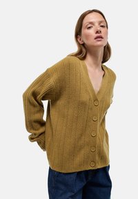 Cardigan côtelé jaune moutarde avec un col en V, cinq boutons marron et des épaules tombantes, associé à un jean taille haute bleu foncé.