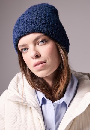 Knit navy blue beanie with a textured pattern, paired with a white puffer jacket. Model has a light blue shirt underneath.