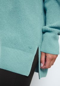 Turquoise knitted sweater with textured pattern, side slits, and ribbed cuffs. Small logo patch on the side adds detail.