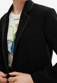 Czarny blezer z białym kontrastowym przeszyciem, klapami z wycięciem i pojedynczą kieszenią na piersi. Nosi się go na białej bluzce z kolorowym kwiatowym wzorem.