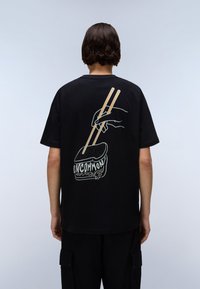 Svart t-skjorte med grafisk design av en hånd som holder spisepinner over mat, med teksten "UNCOMMON" i hvit og beige akenter.