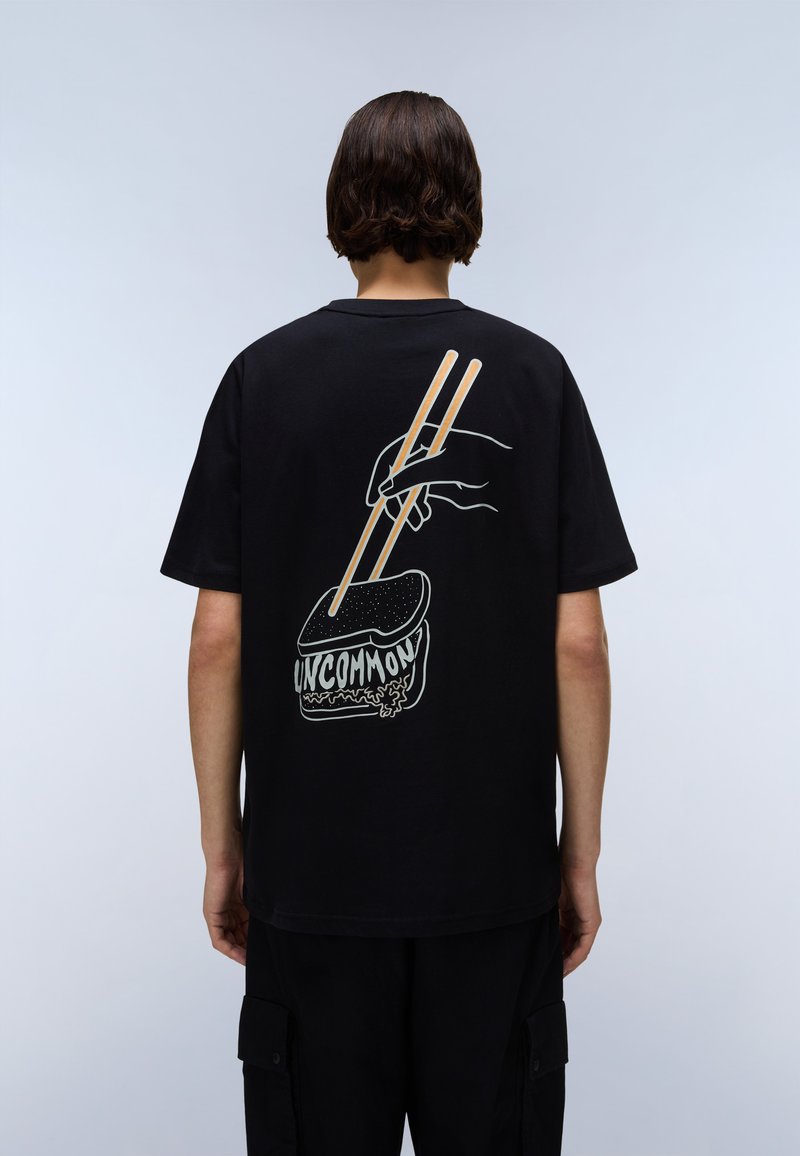 Svart t-skjorte med grafisk design av en hånd som holder spisepinner over mat, med teksten "UNCOMMON" i hvit og beige akenter.