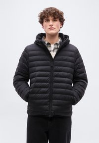 Νέος άνδρας με μπερδεμένα μαλλιά που φοράει μαύρο καπιτονέ μπουφάν με κουκούλα πάνω από ένα καρό πουκάμισο, με τα χέρια στις τσέπες, στέκεται μπροστά από μια απλή φόντο.