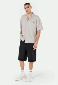 Camisa cinza claro de manga curta com dois bolsos frontais, detalhe do logo preto, combinada com calções de denim pretos, ténis brancos e meias.