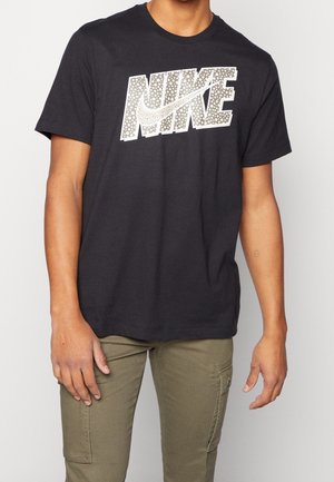 Henkilö, jolla on musta Nike-paita, jossa on pistekuvioinen logo, ja oliivinvihreät cargo-housut, seisoo yksinkertaisen vaalean taustan edessä.