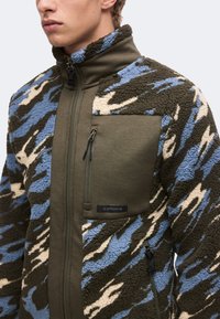 Jeune homme portant une veste polaire zippée avec un motif camouflage bleu, vert et beige et une poche poitrine vert olive avec une fermeture éclair.
