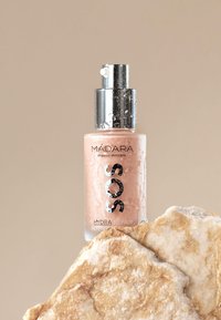 MÁDARA SOS Hydra Repair-serum i en klar glasflaska med silverpump, med en rosa geléliknande konsistens. Ställd på en texturerad klippåta.