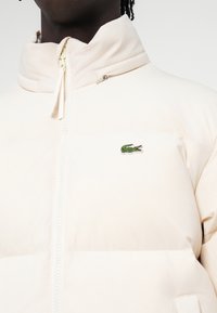 Gaišas beiges puffer jaka ar augstu apkakli un priekšējo rāvējslēdzēju, akcentēta ar mazu zaļu Lacoste krokodila logotipu uz krūtīm.