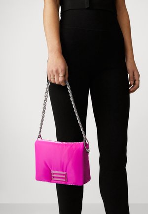 Pink padded handbag with a shiny silver braided strap, featuring a minimalist metal accent on the front, held beside black leggings.