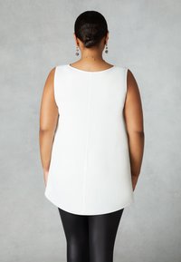 Vit ärmlös topp med rundad fåll och en slät textur, matchad med glänsande svarta leggings. Subtil sömdetalj på baksidan.