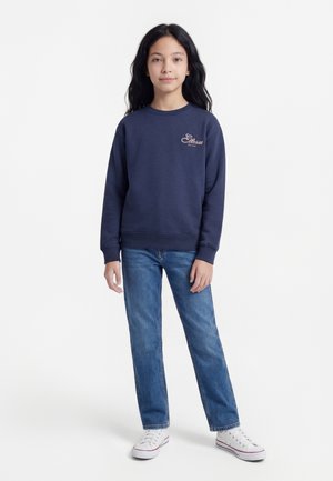 Κορίτσι με μακριά σκούρα μαλλιά που φοράει μπλε φούτερ, μπλε τζιν και λευκά αθλητικά παπούτσια, στέκεται μπροστά σε έναν εντελώς λευκό φόντο.