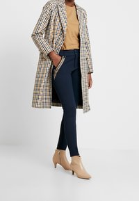 Beige och marinrandig lång kappa över en tan topp, marinblå figurnära byxor med dragkedjedetaljer och beige stövletter med medelhögt klack.