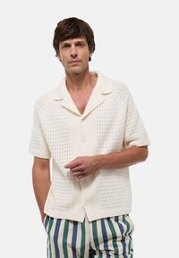 Chemise tricotée couleur crème avec un col pointu, des manches courtes et une fermeture à boutons, présentant un motif texturé. Associée à un pantalon rayé.