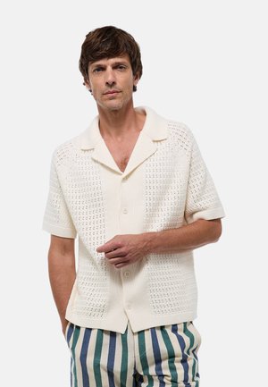 Chemise tricotée couleur crème avec un col pointu, des manches courtes et une fermeture à boutons, présentant un motif texturé. Associée à un pantalon rayé.