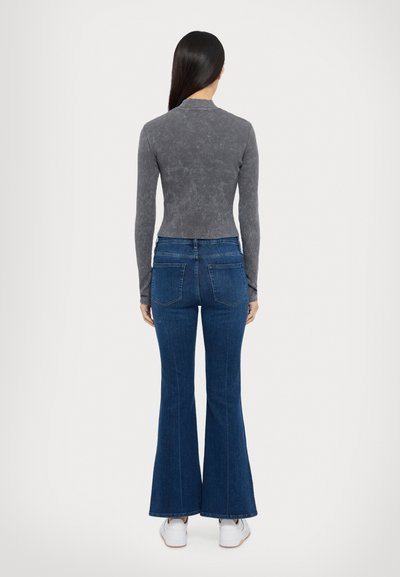 Grå långärmad topp som sitter tajt med hög hals, kombinerad med blå utsvängda jeans och vita sneakers, sedd bakifrån.