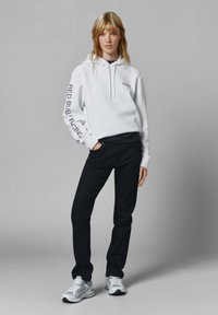 Biely hoodie s šnúrkou, na rukáve potlač "Red Bull Racing"; skombinovaný s čiernymi džínsami a šedými teniskami. Hladká tkanina, uvoľnený strih.