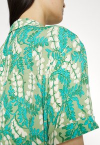 Grüne Stoffbluse mit einem floralen Muster und türkisfarbenen sowie weißen Akzenten, kurzen Ärmeln und einem klassischen Kragenstil.