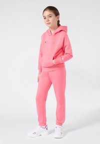 Ragazza giovane in piedi, che indossa un felpa rosa Champion e pantaloni della tuta abbinati, con scarpe bianche e viola, su uno sfondo bianco.