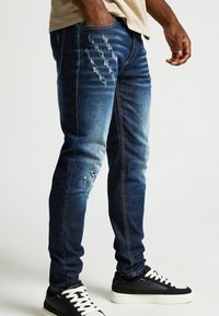 Zila denima skinny džinsi ar nolietotām detaļām, vieglu izbalēšanu, piecām kabatām un ar rāvējslēdzēju, savienoti ar melnbaltiem čībām.