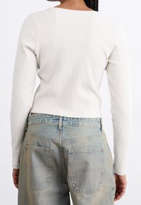 Top blanco de manga larga con canalé, combinado con jeans de tiro alto de mezclilla azul claro. Las características notables incluyen un tejido texturizado y costuras visibles en los bolsillos.