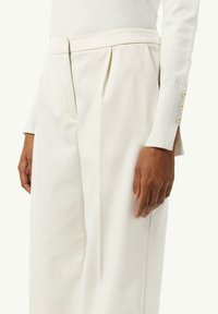 White tailored trousers with pleats, featuring a high waistline and side pockets. The fabric has a smooth texture, complemented by a fitted cream top.