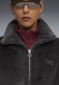 Gray faux fur jacket with a high collar and maroon inner layer, featuring a silver zipper and a logo emblem, paired with brown tinted sunglasses.