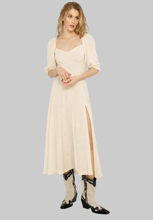 Donna con capelli biondi mossi che indossa un abito midi bianco sporco con maniche a sbuffo e spacco alto sulla coscia, abbinato a stivali cowboy neri e beige.