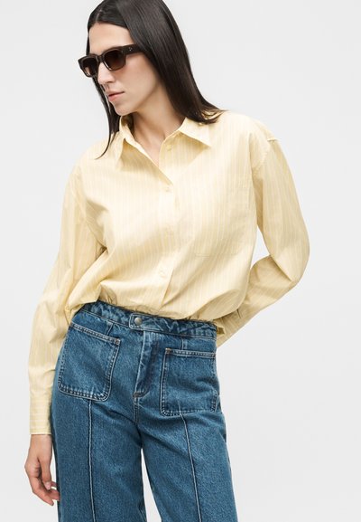 Γυναίκα που φοράει σκούρα γυαλιά ηλίου, ανοιχτό κίτρινο ριγέ πουκάμισο με κουμπιά και ψηλόμεσο τζιν μπλε με εμπρόσθιες τσέπες με πάτσες.