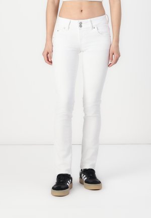 Frau trägt weiße Slim-Fit-Jeans und schwarze Sneakers mit weißen Streifen, steht vor einem schlichten weißen Hintergrund.