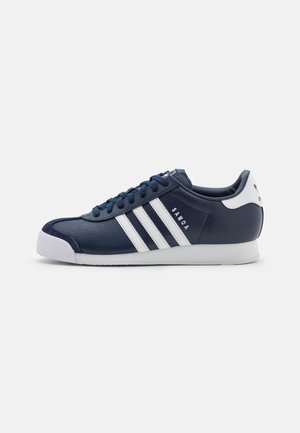 Granatowy sneaker Adidas Samoa z białymi paskami, białą zakładką na pięcie, sznurowanym designem i teksturowanym czubkiem, na jednolitym jasnym tle.