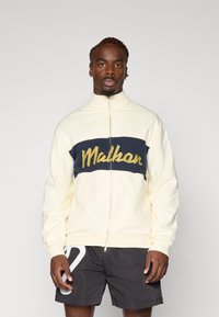 Cremefarvet zip-up sweatshirt med navy vandret panel, der har gul tekst "Malhar" på tværs af brystet. Glat stof og afslappet pasform.
