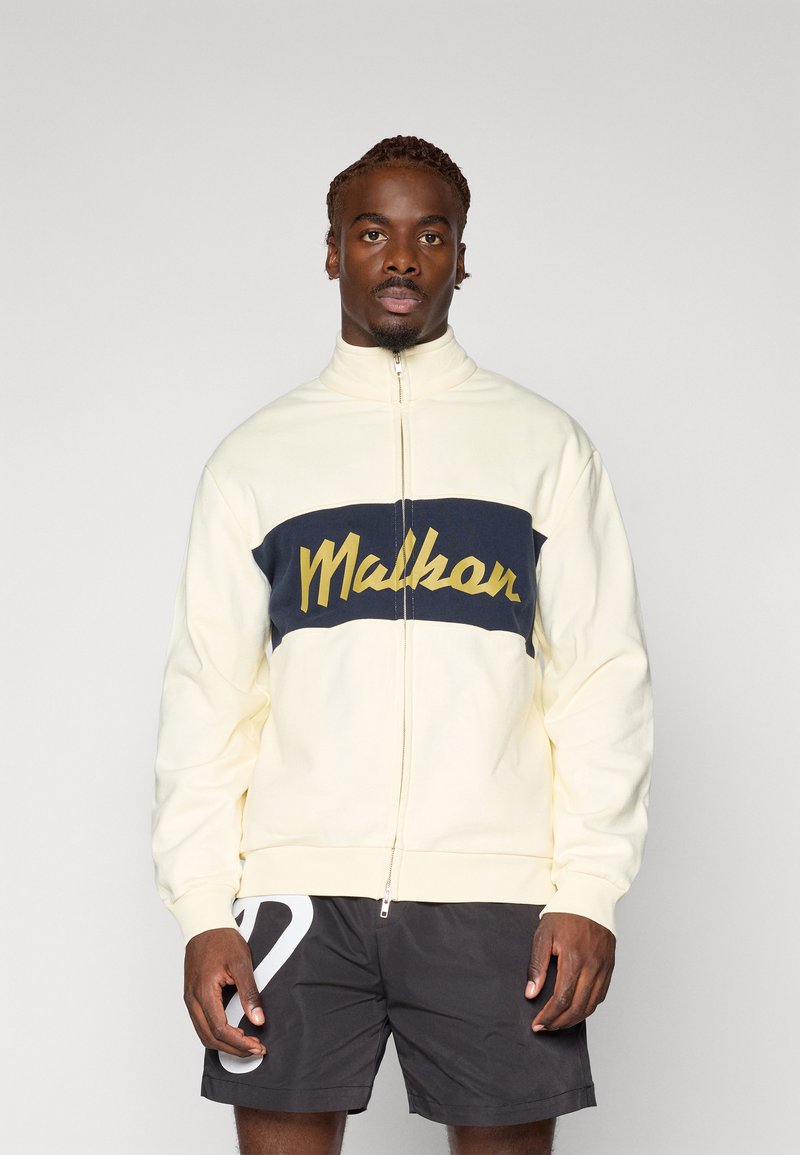 Cremefarvet zip-up sweatshirt med navy vandret panel, der har gul tekst "Malhar" på tværs af brystet. Glat stof og afslappet pasform.