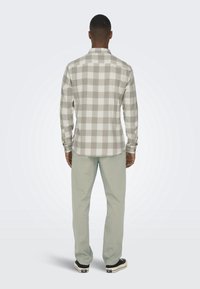 Šedá a bílá kostkovaná košile na knoflíky s dlouhými rukávy, s límečkem a zaobleným lemem, doplněná o světle zelené kalhoty a černé tenisky.