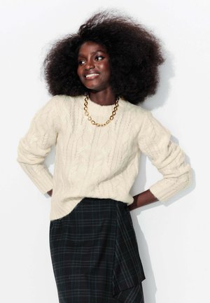 Pull en crème à mailles, avec des poignets côtelés, associé à une jupe plissée à carreaux noirs. Un collier en chaîne dorée remarquable accentue le look.