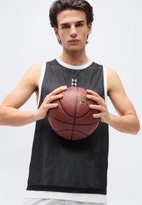 Νέος άνδρας με μαύρη αμάνικη αθλητική φανέλα κρατάει ένα μπάσκετ και με τα δύο χέρια στο ύψος του στήθους, κοιτάζοντας μπροστά.