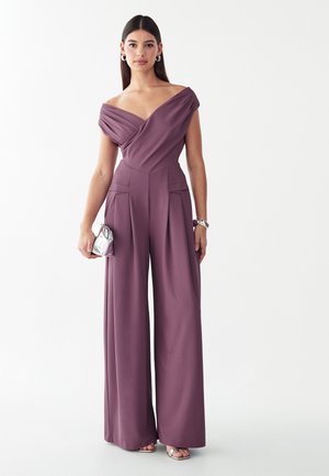 Woman wearing a mauve off-shoulder jumpsuit with wide legs, holding a small silver clutch bag, against a plain white background.
